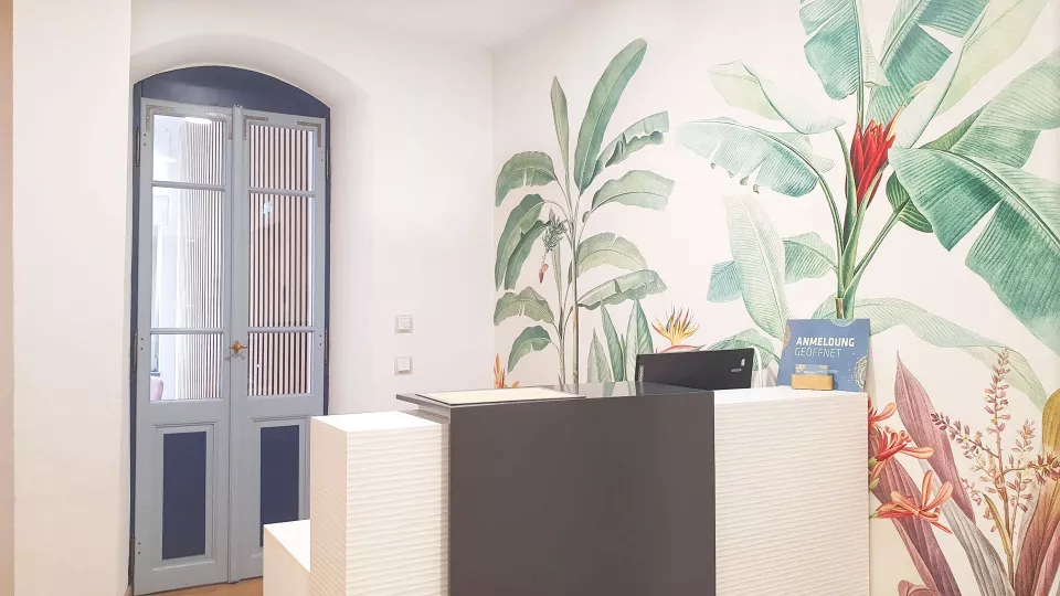 Modern gestalteter Empfangsbereich der Praxis Seeger & Tändler mit Anmeldungstheke, tropischer Pflanzenwand und Eingangstür für eine einladende Atmosphäre.